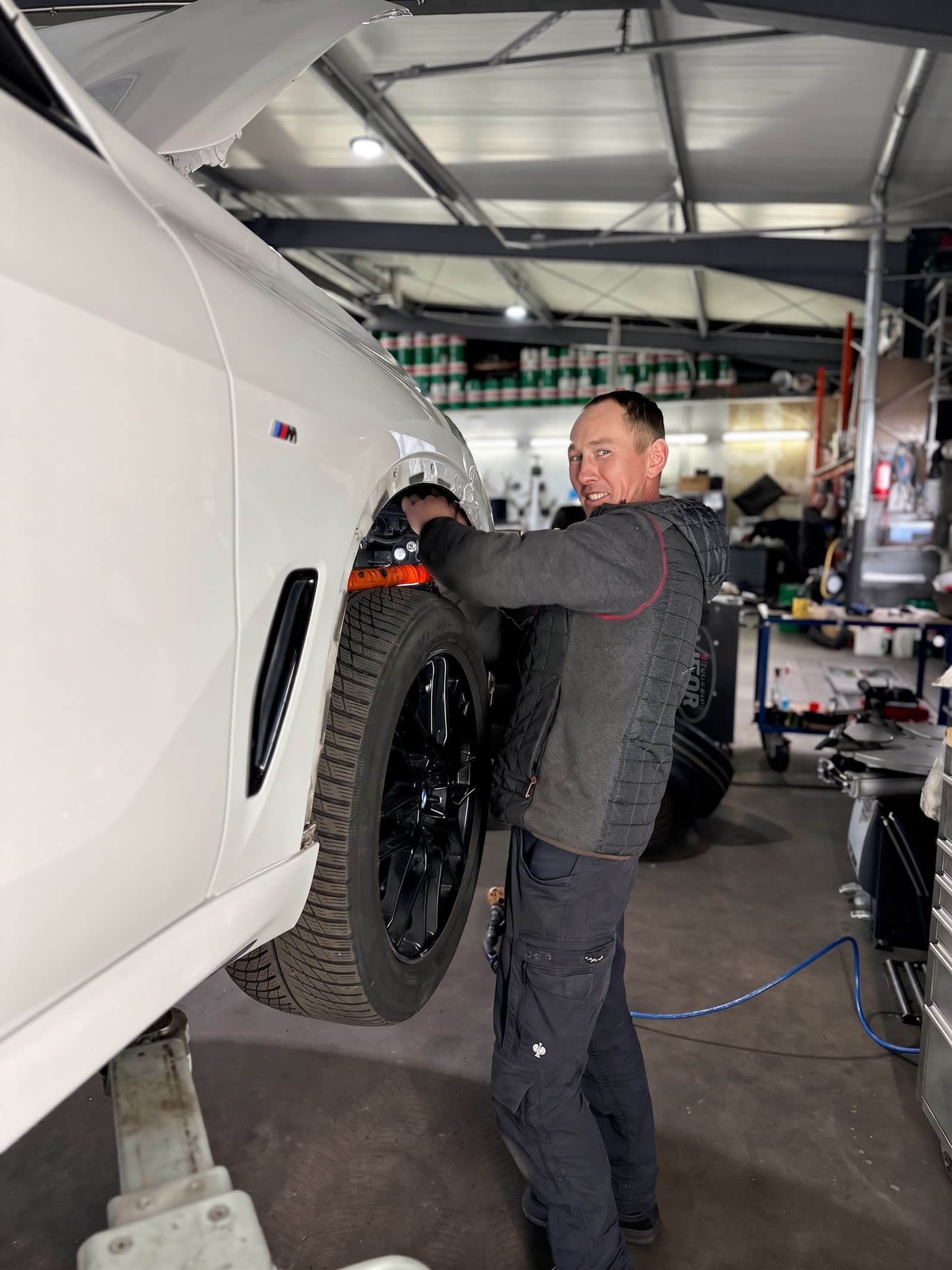 StarWash-Mechaniker lächelt am Vorderrad eines weißen BMW M, M-Logo am Bremssattel sichtbar